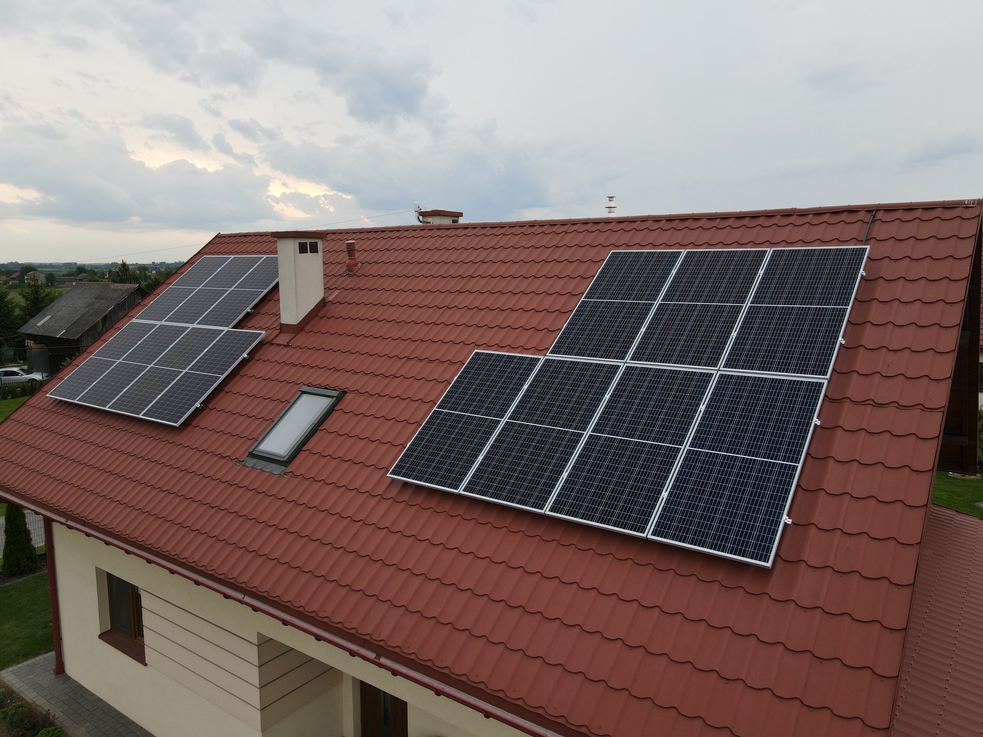 Widok z góry na dom z czerwonym dachem i zamontowanymi panelami słonecznymi, widoczne okno dachowe i komin.