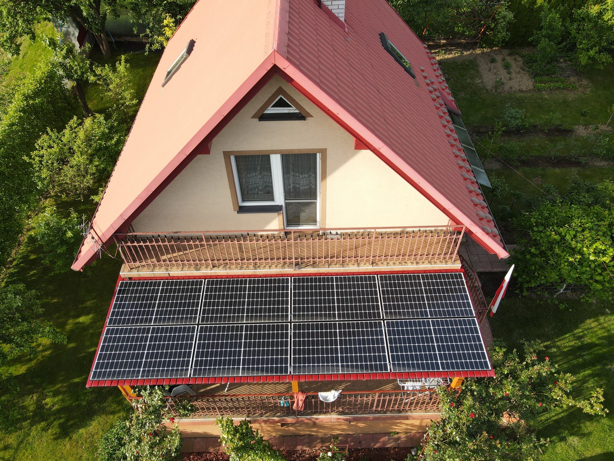 Widok z góry na dom z czerwonym dachem i panelami fotowoltaicznymi zamontowanymi na balkonie, obok powiewa flaga Polski.