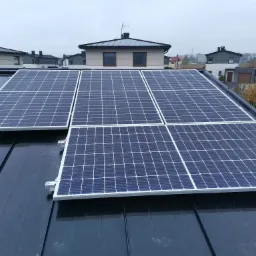 Instalacja paneli słonecznych na dachu domu jednorodzinnego, widoczne moduły w układzie prostokątnym, tło stanowią inne domy w zabudowie szeregowej i pochmurne niebo.