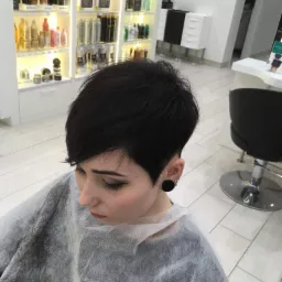 Kobieta z krótką, czarną fryzurą pixie cut, siedząca w salonie z ochronnym okryciem na ramionach, widoczny kolczyk typu tunel w uchu, na tle regału z kosmetykami do włosów i białych mebli.