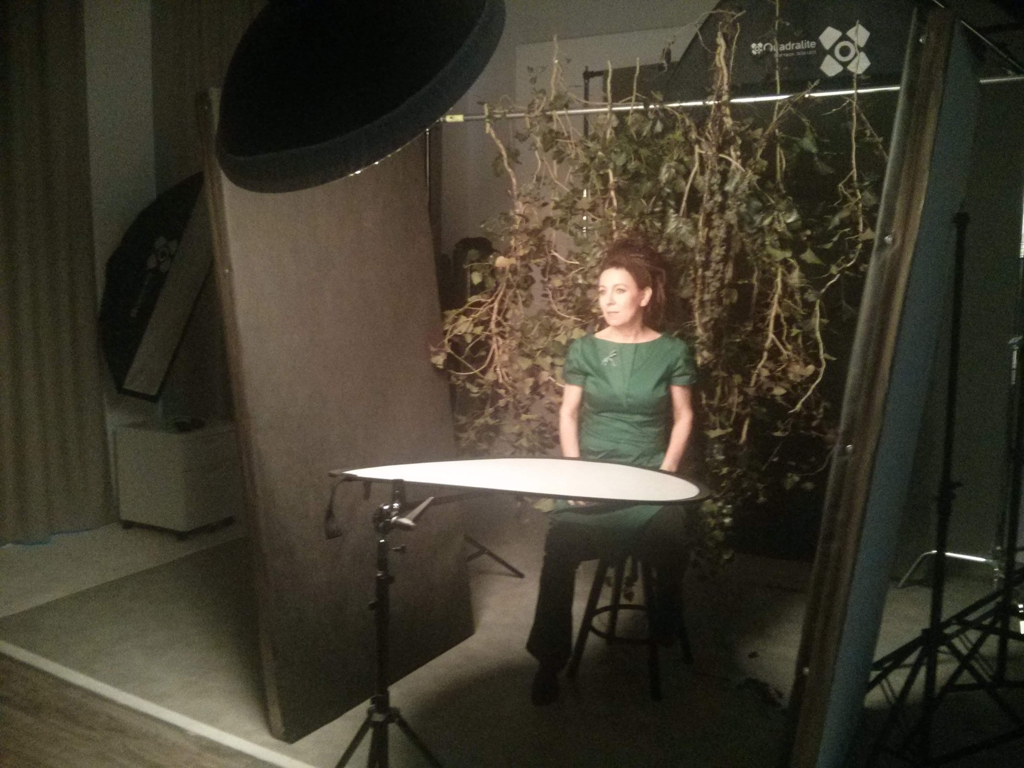Kobieta w zielonej sukience siedzi na stołku w studio fotograficznym z dekoracjami z bluszczu, oświetleniem i blendami.