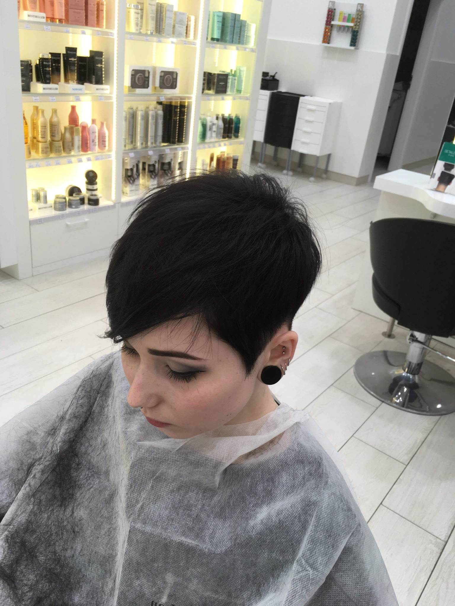 Kobieta z krótką, czarną fryzurą pixie cut, siedząca w salonie z ochronnym okryciem na ramionach, widoczny kolczyk typu tunel w uchu, na tle regału z kosmetykami do włosów i białych mebli.
