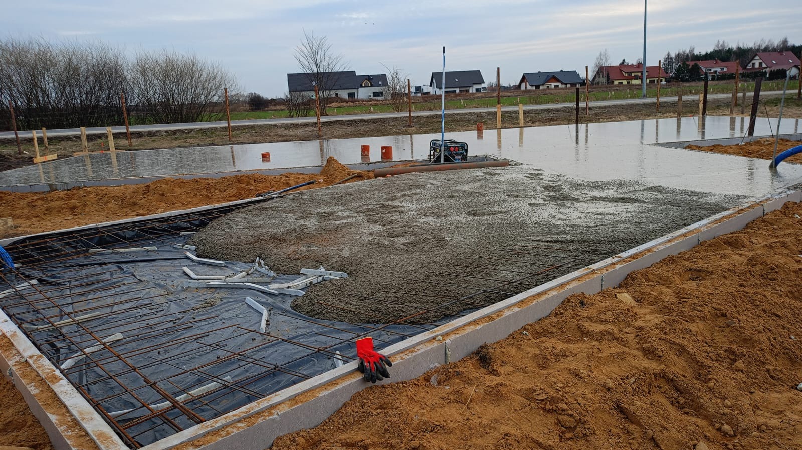 Wylewanie betonu na zbrojenie płyty fundamentowej w szalunku. Widoczne rękawice robocze, pompa do betonu i częściowo zalana powierzchnia. Tło: zabudowa jednorodzinna.
