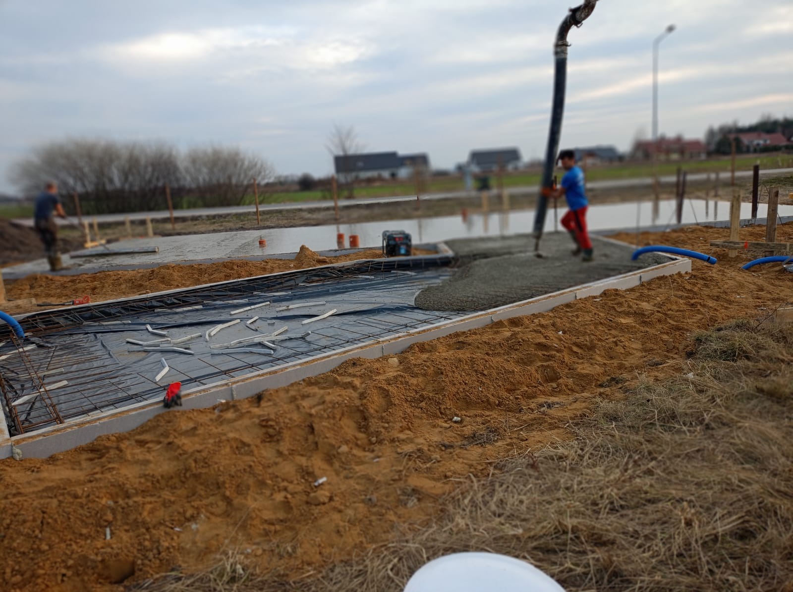Wylewanie betonu na przygotowaną płytę fundamentową zbrojoną stalą i folią, widoczne szalunki, pracownik z pompą do betonu. Prace budowlane w Dobrzykowicach.