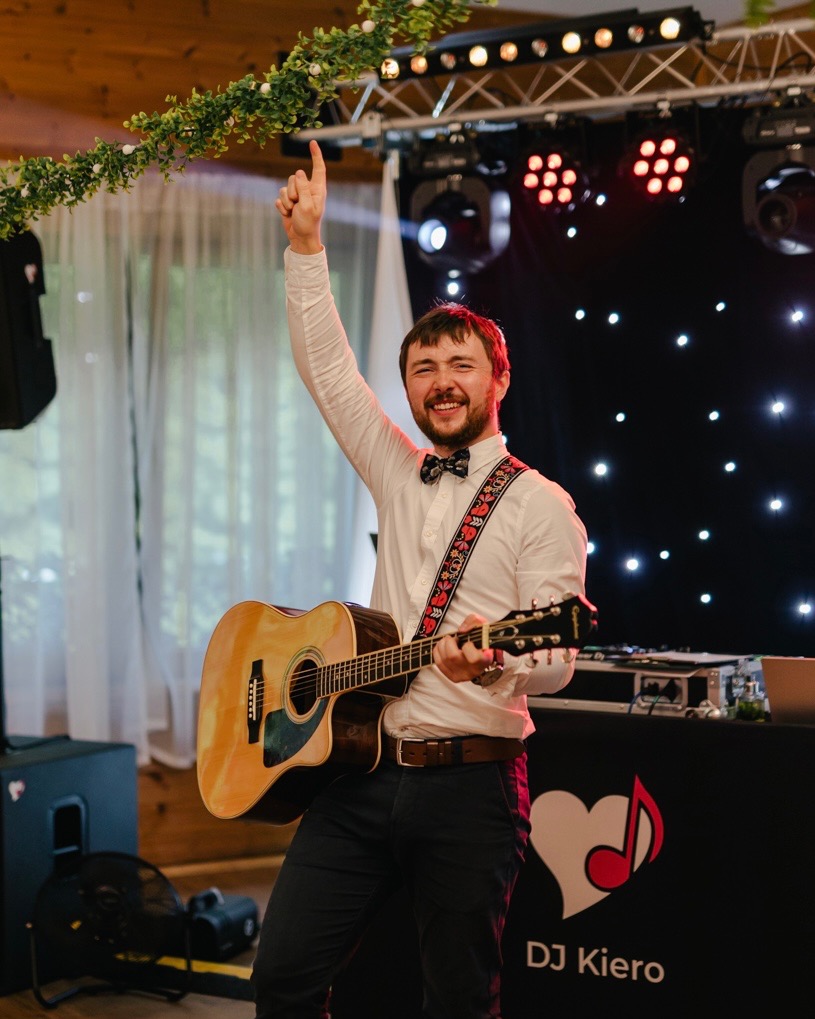 Uśmiechnięty muzyk z gitarą akustyczną, w muszce i z uniesionym palcem, na tle sceny z oświetleniem i logo DJ-a.