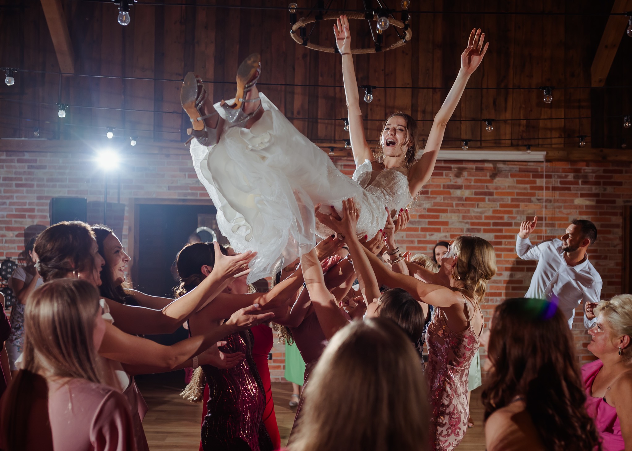 Uśmiechnięta panna młoda w białej sukni uniesiona w górę przez gości weselnych na tle ceglanej ściany i drewnianego sufitu z żarówkami