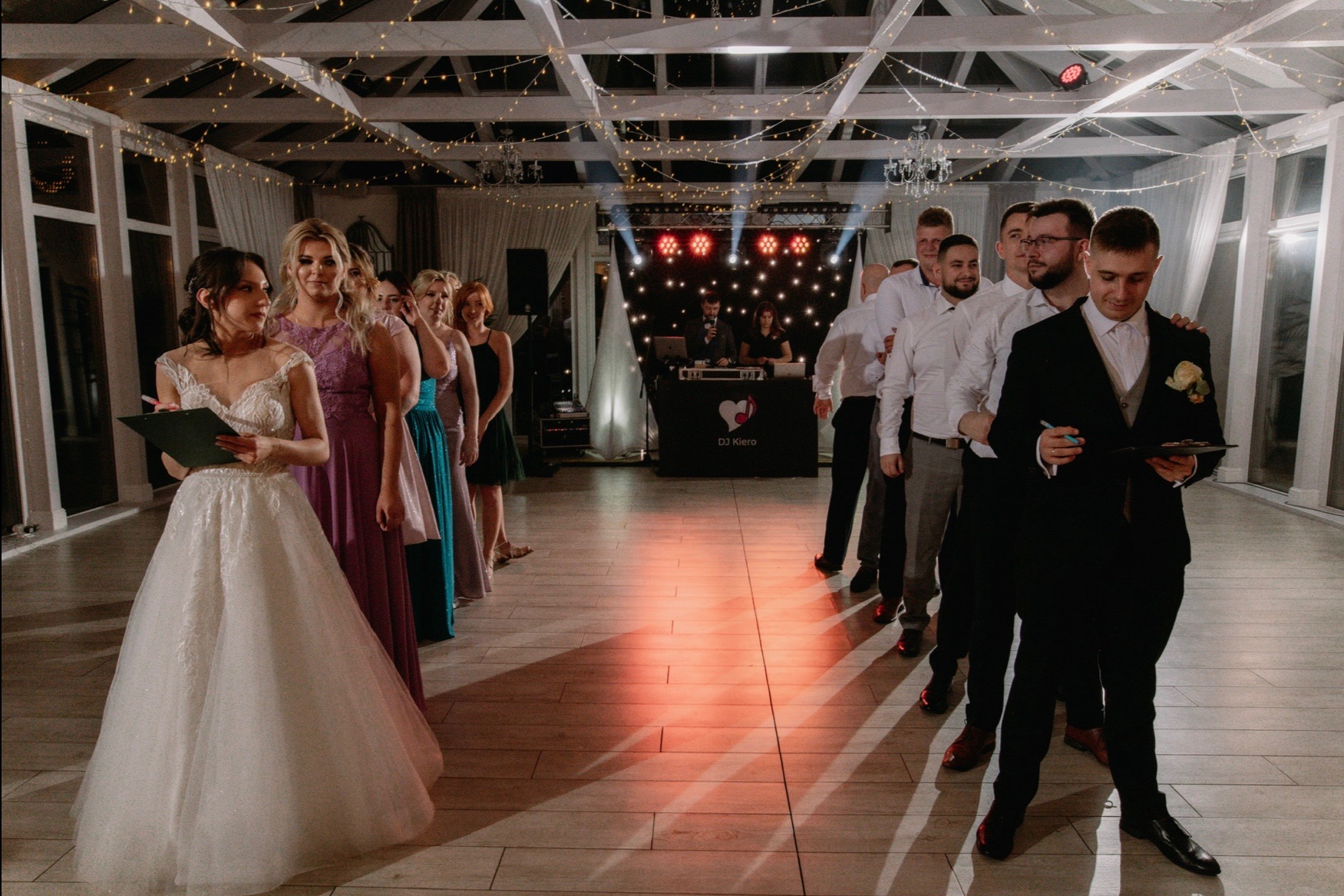 Panna młoda z listą gości i drużba ustawieni w kolejce na sali weselnej, w tle DJ z oświetleniem.