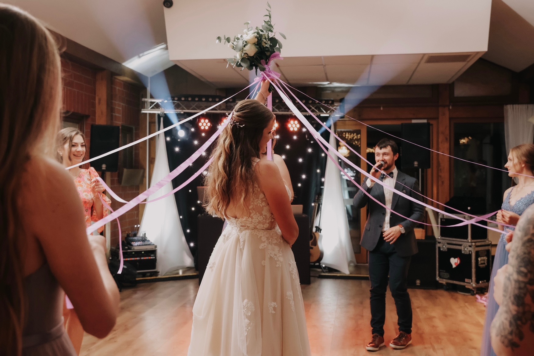 Panna młoda trzyma bukiet podczas wesela, tancerze z różowymi wstążkami, w tle grający wokalista i sprzęt nagłośnieniowy.