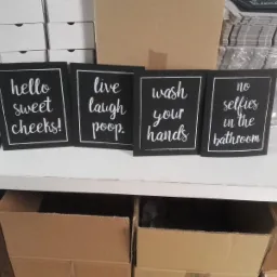 Cztery czarne tabliczki z białymi napisami w ramkach: 'hello sweet cheeks!', 'live laugh poop.', 'wash your hands', 'no selfies in the bathroom', ustawione na białej półce, za nimi pudełka...