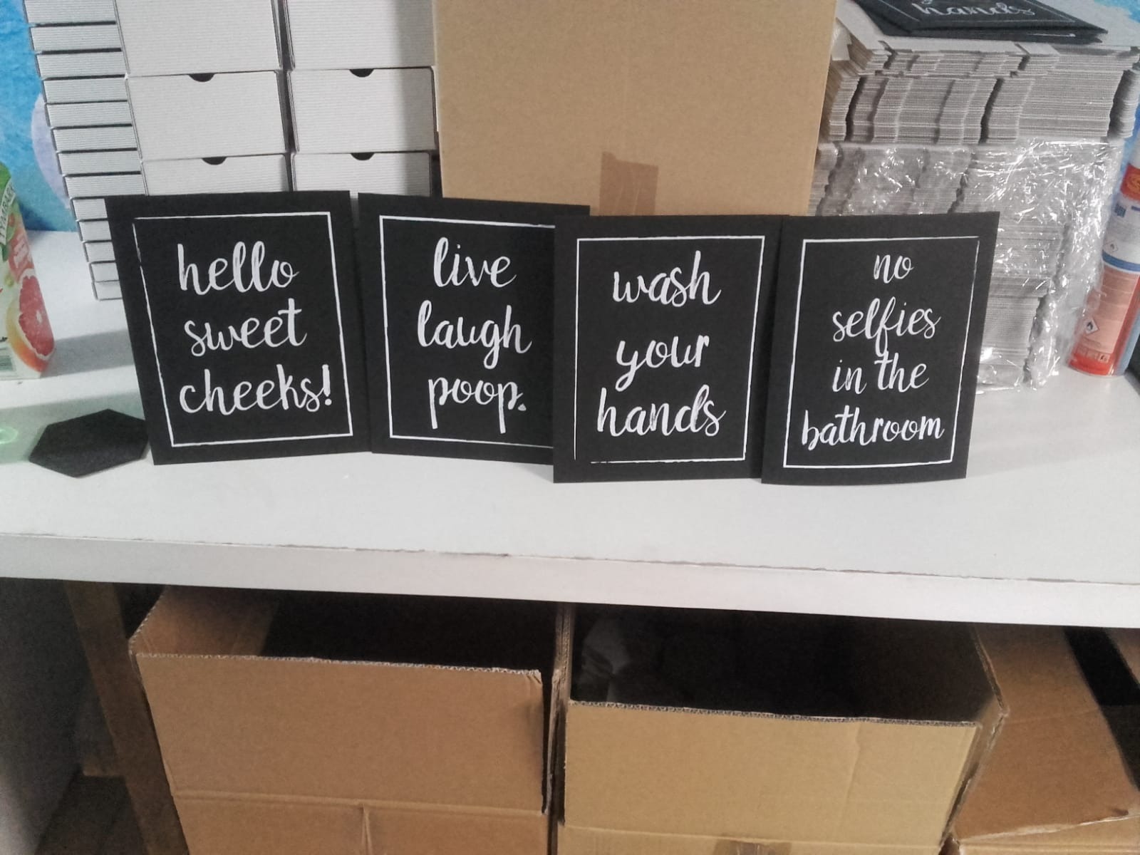 Cztery czarne tabliczki z białymi napisami w ramkach: 'hello sweet cheeks!', 'live laugh poop.', 'wash your hands', 'no selfies in the bathroom', ustawione na białej półce, za nimi pudełka...