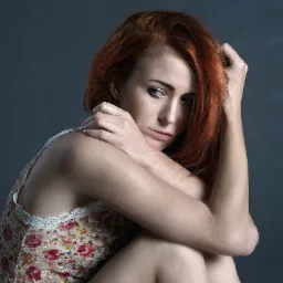 Portret kobiety o rudych włosach, zamyślonym spojrzeniu i ramionach obejmujących kolana, na ciemnym tle.