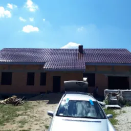 Dom w trakcie budowy z nowym, bordowym dachem z dachówki ceramicznej, widoczna drabina oparta o budynek, samochody na pierwszym planie.