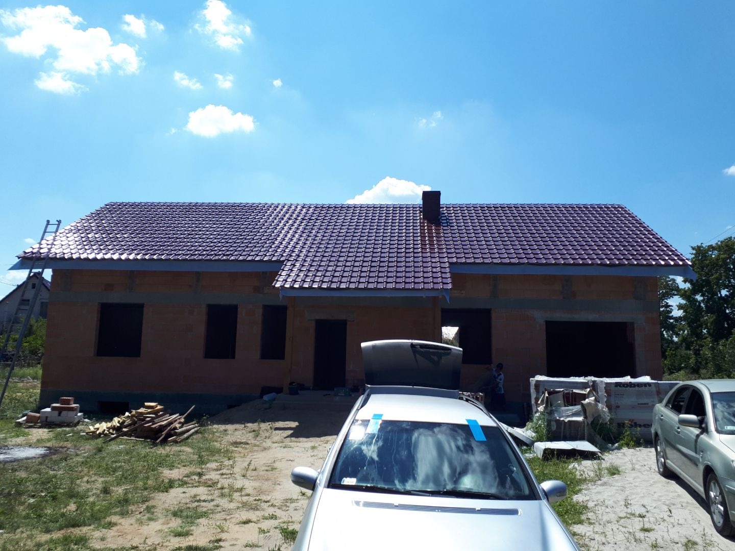 Dom w trakcie budowy z nowym, bordowym dachem z dachówki ceramicznej, widoczna drabina oparta o budynek, samochody na pierwszym planie.