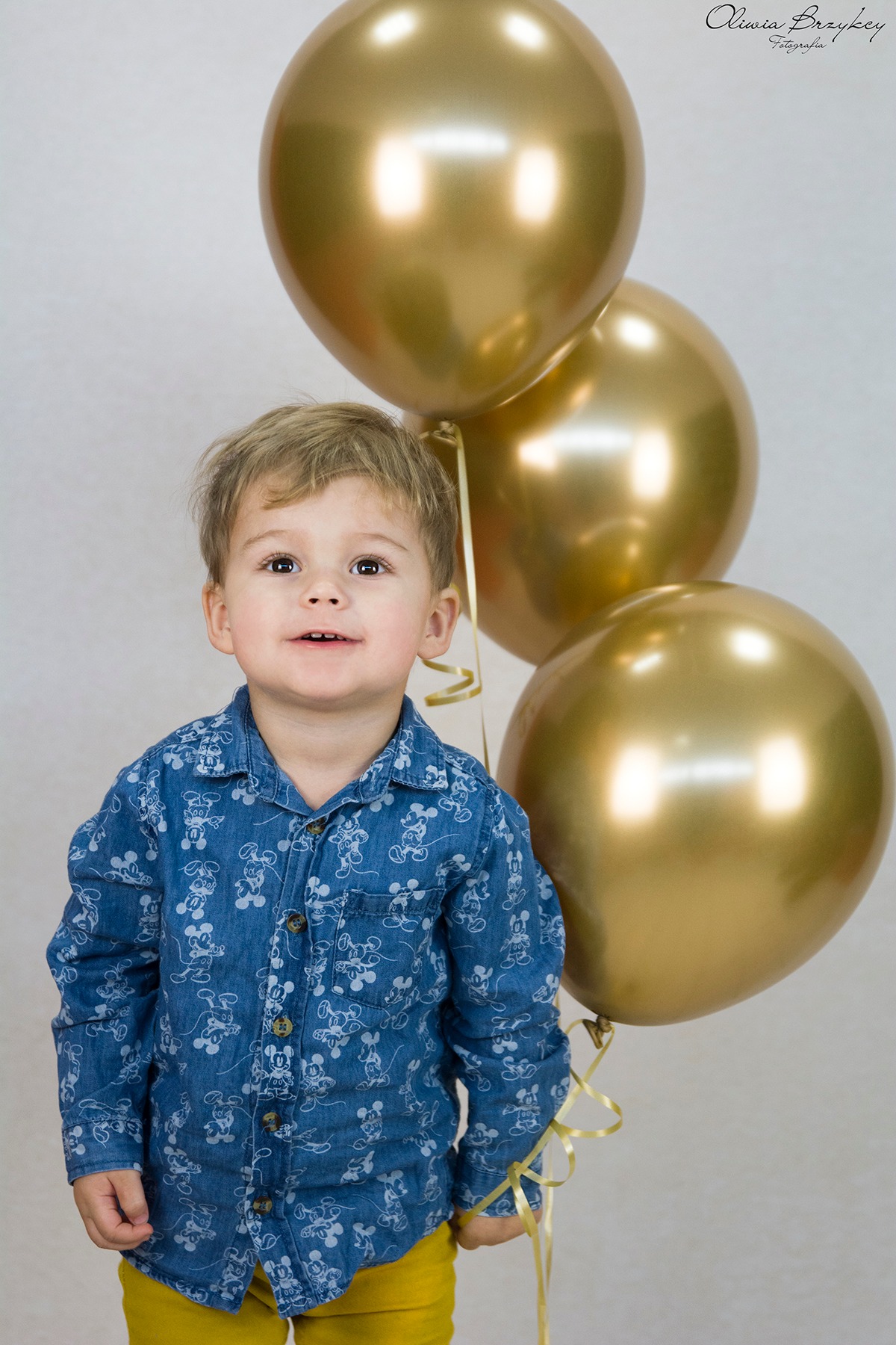 Portret uśmiechniętego chłopca w koszuli w myszki Mickey, z trzema złotymi balonami na sznurkach, na jasnym tle.