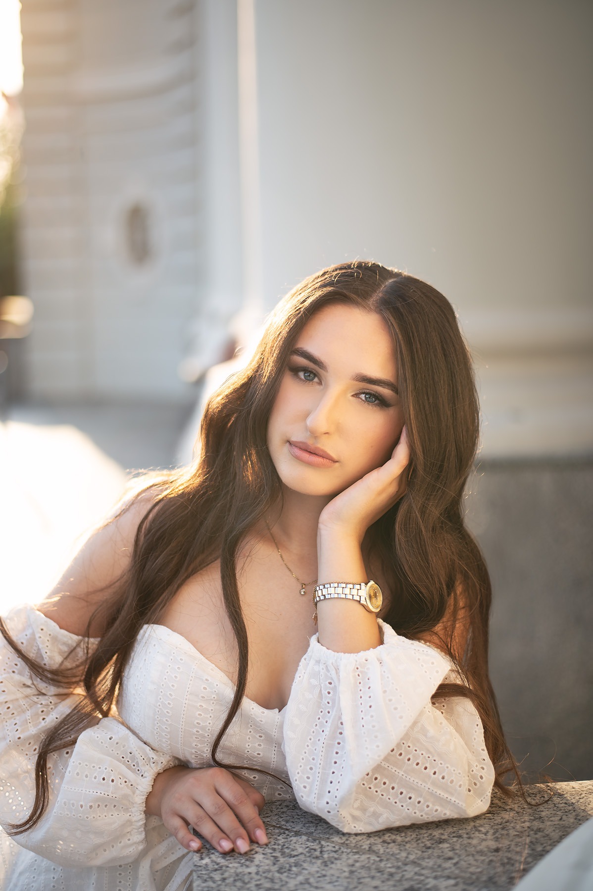 Portret młodej kobiety z długimi brązowymi włosami, w białej sukience z ażurowymi rękawami, opierającej się o kamienny blat, z delikatnym makijażem i złotym zegarkiem.