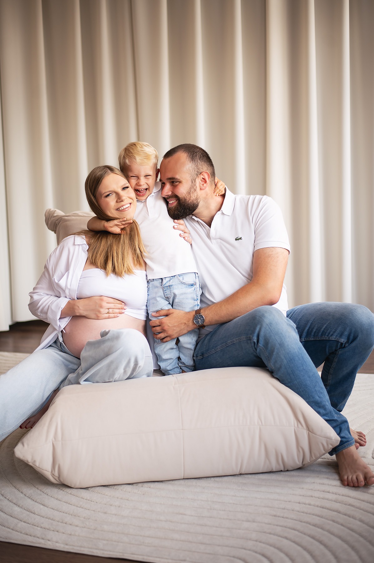 Uśmiechnięta, ciężarna kobieta z mężem i synkiem w jasnym wnętrzu. Rodzinne ujęcie z sesji ciążowej, naturalne światło, beżowe tło, miękki dywan.