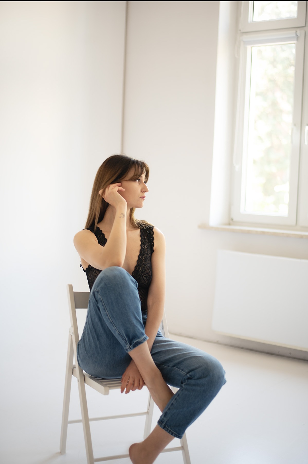 Kobieca sesja w Bydgoszczy: Brunetka w czarnym body i jeansach siedzi na krześle, spoglądając w bok na tle białej ściany i okna. Subtelny tatuaż na ramieniu.