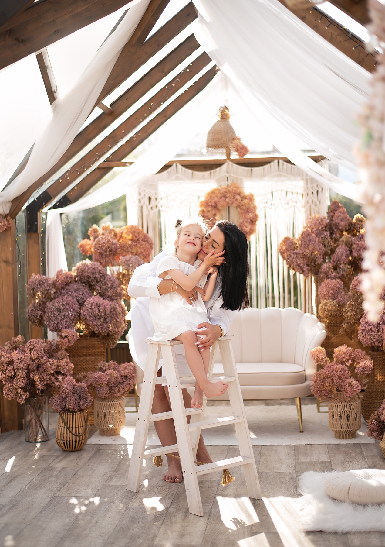Czuły portret matki całującej córkę w policzek, siedzącą na białej drabinie w otoczeniu kwiatowych dekoracji w jasnym, przeszklonym pomieszczeniu.