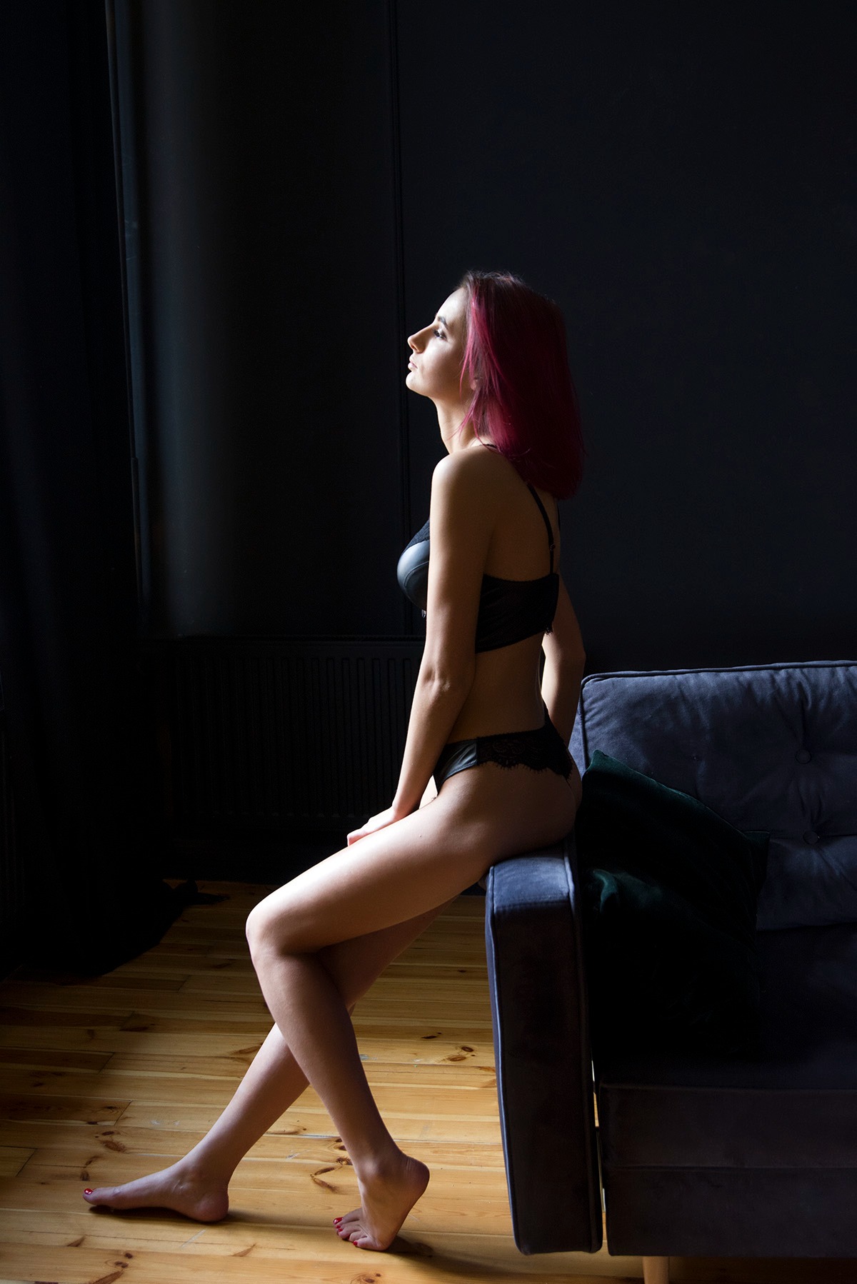 Kobieta w czarnej bieliźnie siedzi na sofie na tle ciemnej ściany. Ujęcie z profilu, podkreślające sylwetkę i subtelne oświetlenie. Widoczne drewniane panele podłogowe.