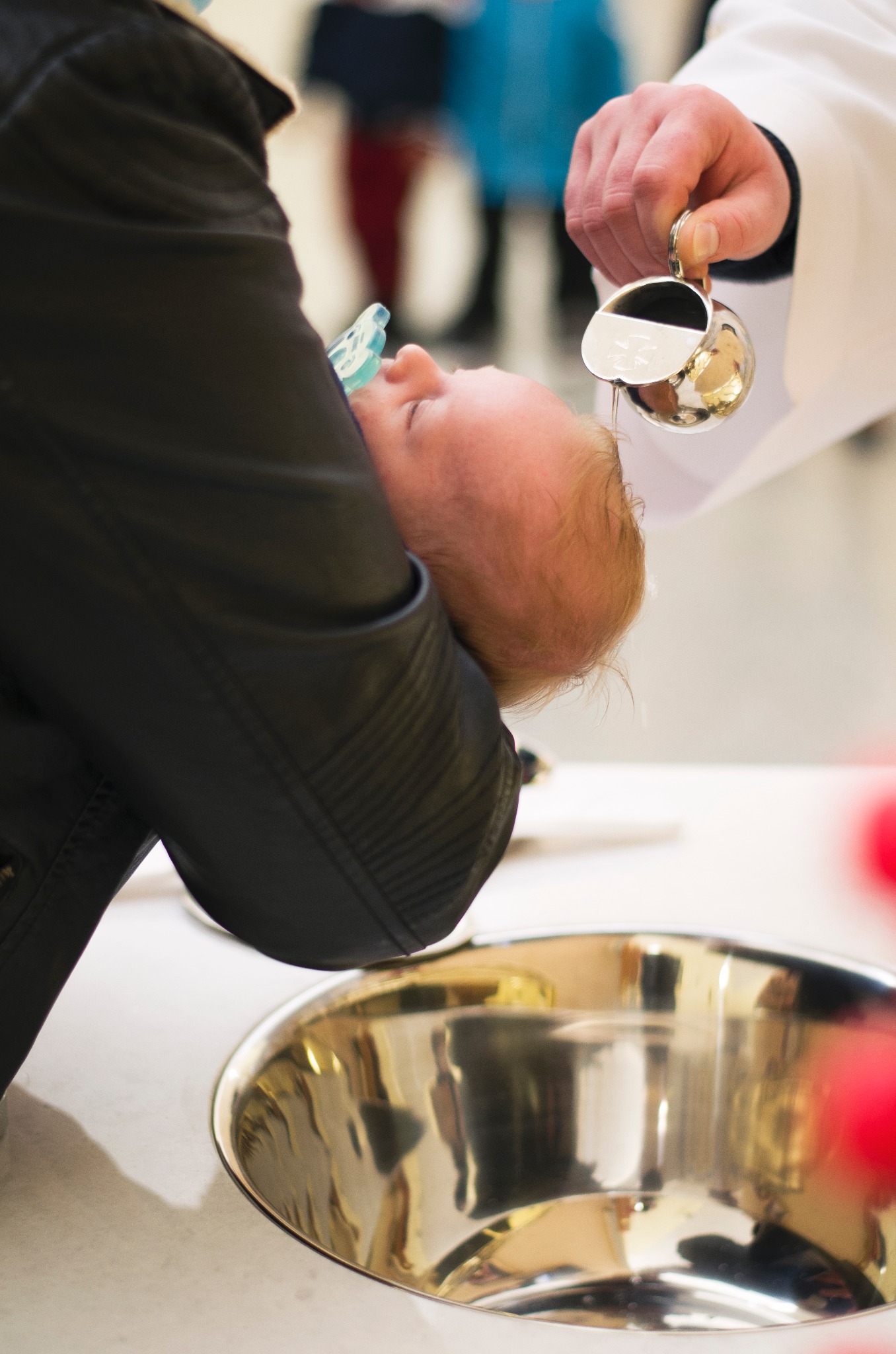 Kąpiel noworodka podczas ceremonii: Woda z dzbanka polewana na głowę dziecka trzymanego przez osobę w czarnej kurtce, srebrna miska poniżej.