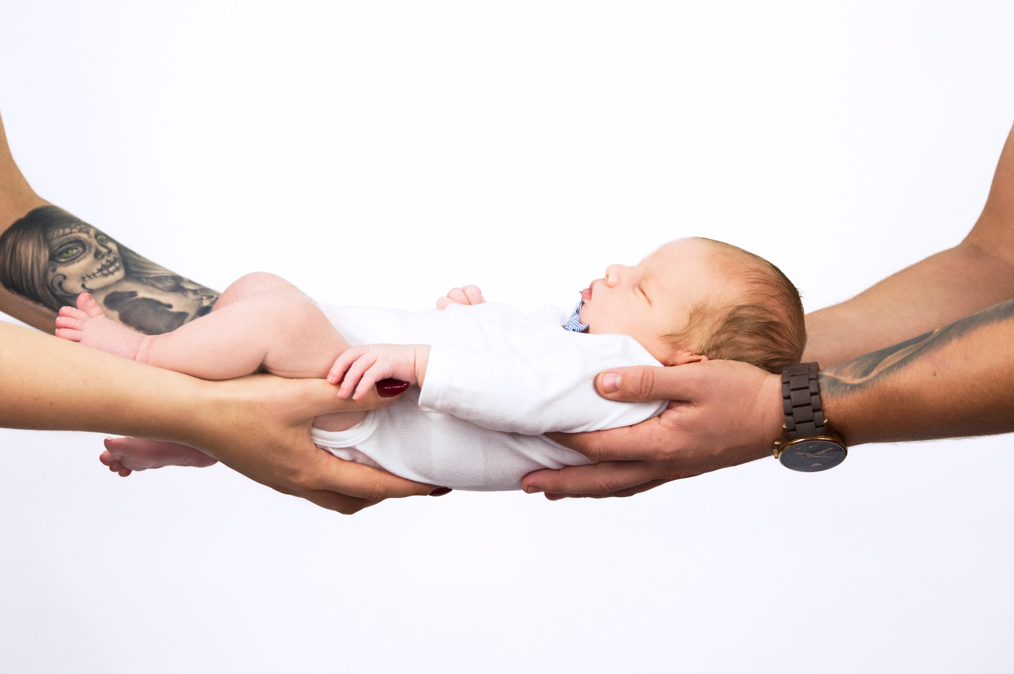 Noworodek w białym body, trzymany w dłoniach rodziców z widocznymi tatuażami i zegarkiem na ręce, na białym tle.