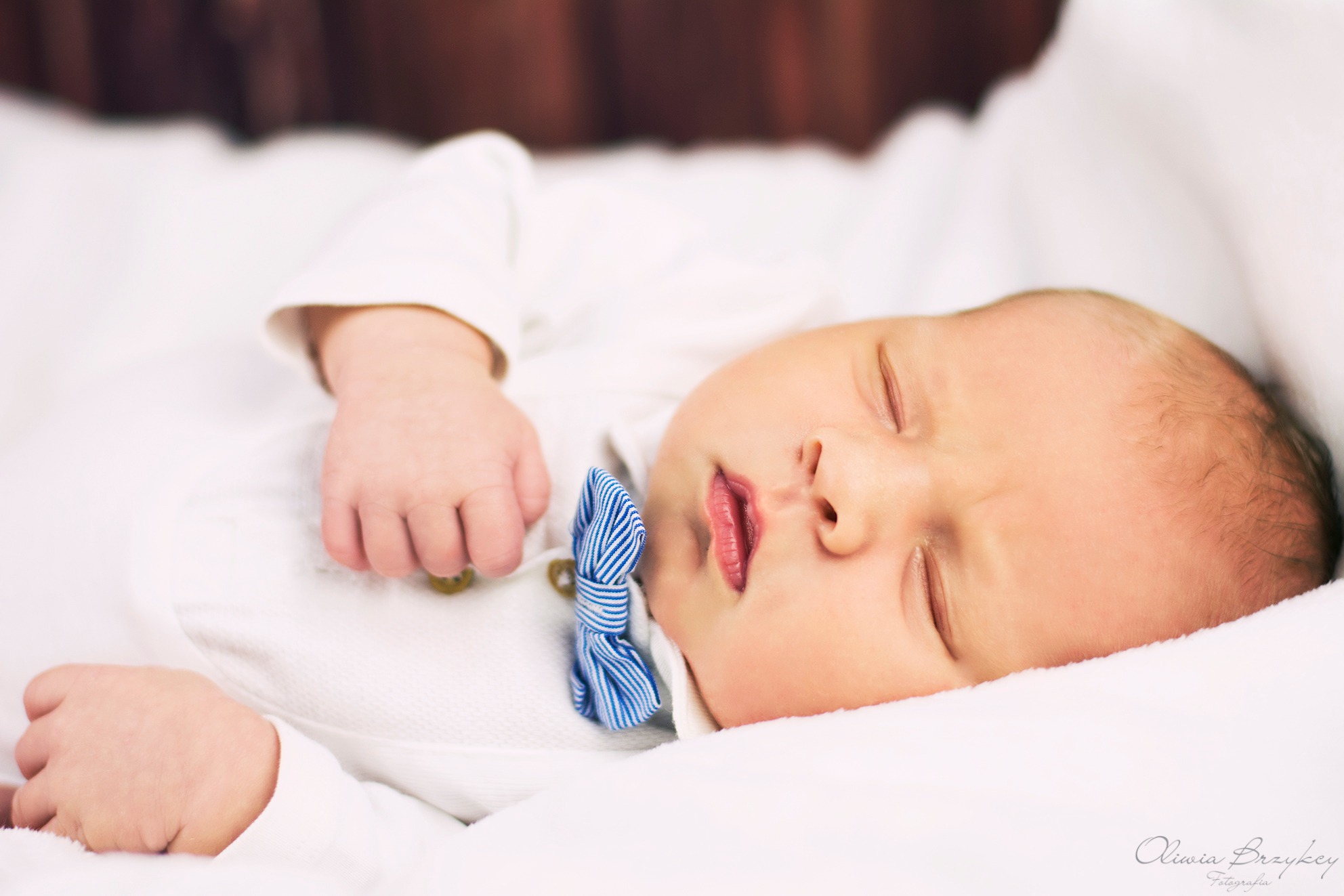 Śpiący noworodek w białym ubranku z niebieską, pasiastą muchą, leżący na białym kocyku.