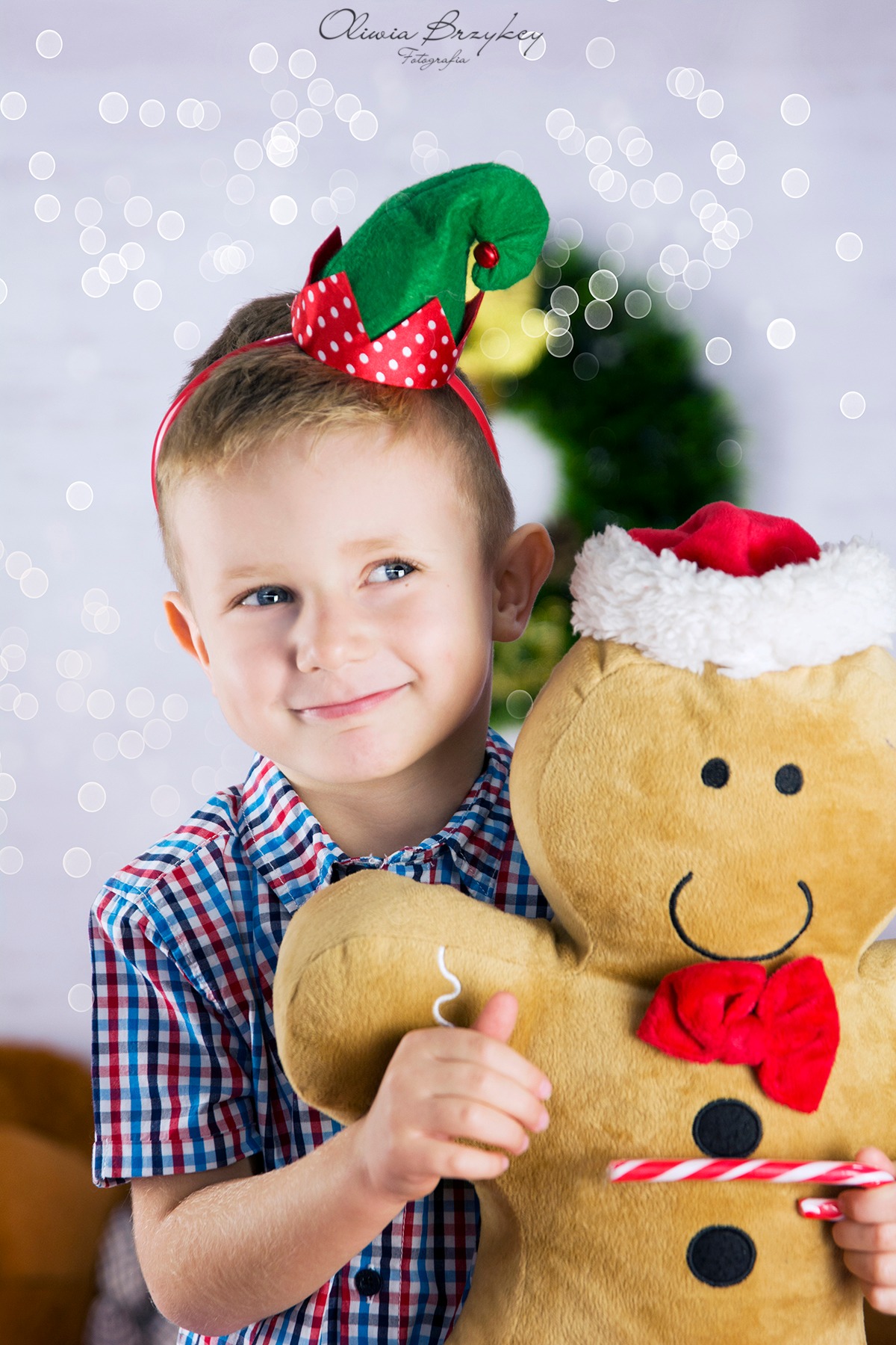Uśmiechnięty chłopiec w czapce elfa i kraciastej koszuli przytula pluszowego piernika z czerwoną muchą i laską cukrową na tle białych światełek.