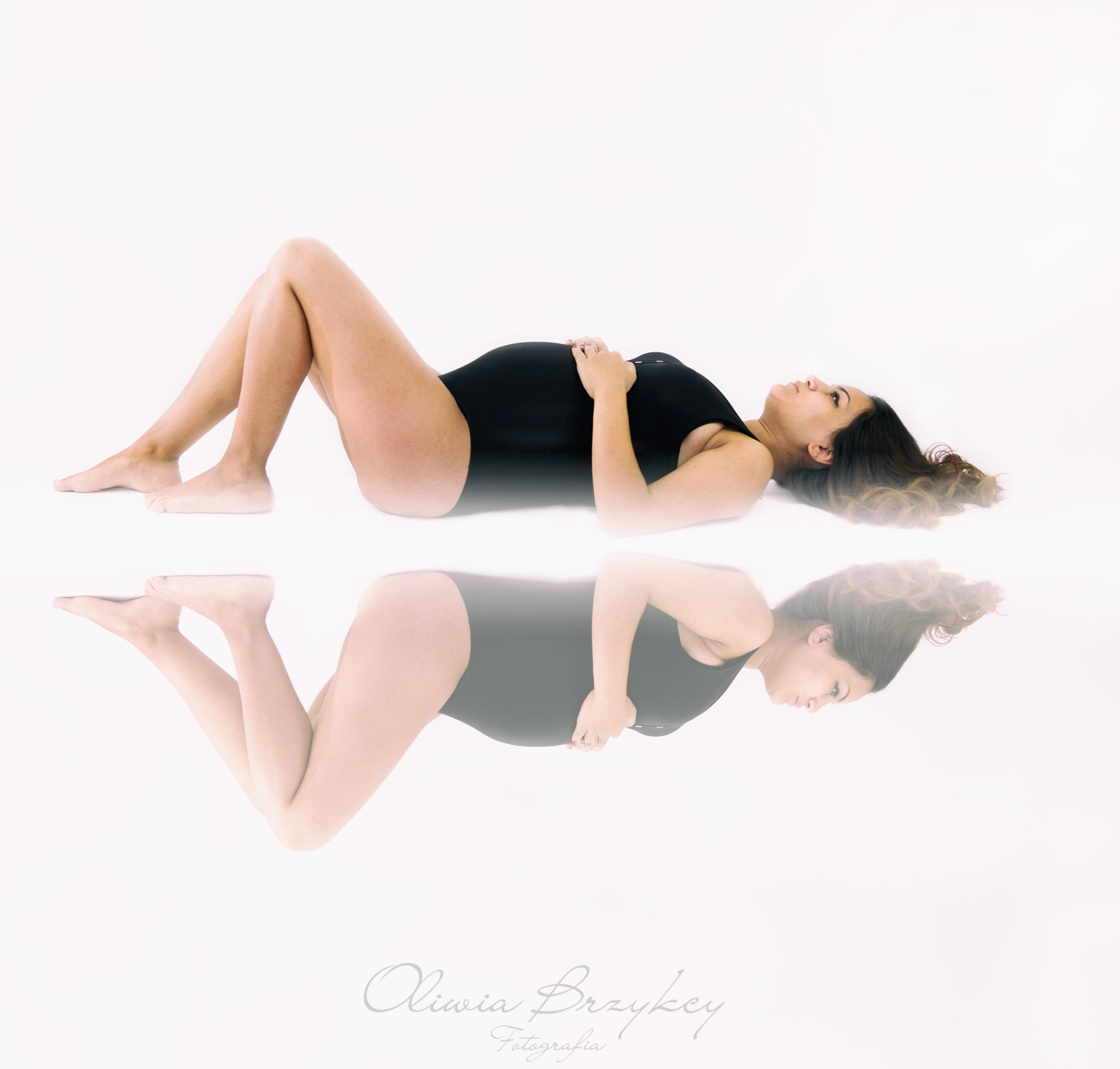 Kobieta w czarnym stroju kąpielowym leży na białym tle, trzymając dłonie na brzuchu; odbicie poniżej. Jasne, studyjne oświetlenie.