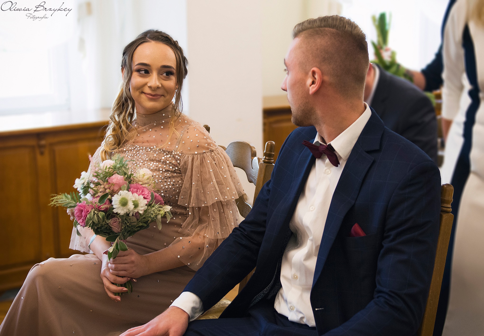 Panna młoda w beżowej sukni z bukietem kwiatów spogląda na pana młodego w granatowym garniturze z bordową muszką, siedzących na drewnianych krzesłach.