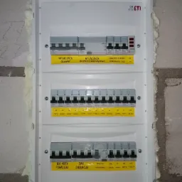 Rozdzielnica elektryczna z wyłącznikami i zabezpieczeniami, zamontowana na surowej ścianie z pianką montażową wokół obudowy, widoczne żółte etykiety z opisami obwodów.