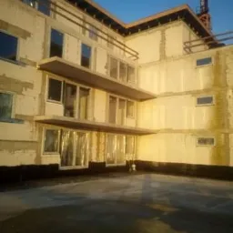 Budynek w trakcie budowy z widocznymi oknami, niektóre okna zakryte folią, na tle błękitnego nieba z widocznym żurawiem budowlanym.