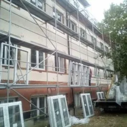 Wymiana okien w budynku wielorodzinnym z użyciem rusztowania, okna częściowo zamontowane, część przygotowana do montażu, widoczny samochód dostawczy.