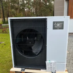 Biała jednostka zewnętrzna pompy ciepła Mitsubishi Zubadan R32 na drewnianej palecie, na trawniku przed domem.