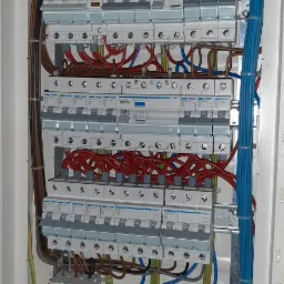 Otwarta skrzynka bezpiecznikowa Hager z widocznymi bezpiecznikami, przewodami w kolorach niebieskim, czerwonym, brązowym i zielono-żółtym, starannie ułożonymi wewnątrz obudowy.