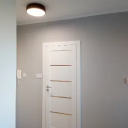 Wnętrze mieszkania po remoncie: białe drzwi wewnętrzne z poziomymi przeszkleniami, szara ściana z tapetą w pionowe prążki, okrągła lampa sufitowa i biały domofon.