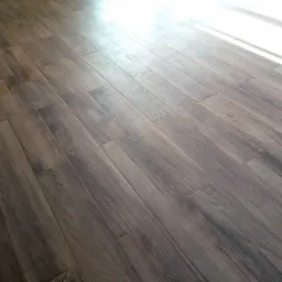 Ułożone panele podłogowe w odcieniach brązu z widocznym śladem/plamą w lewej części kadru, oświetlone naturalnym światłem wpadającym z góry po prawej stronie.