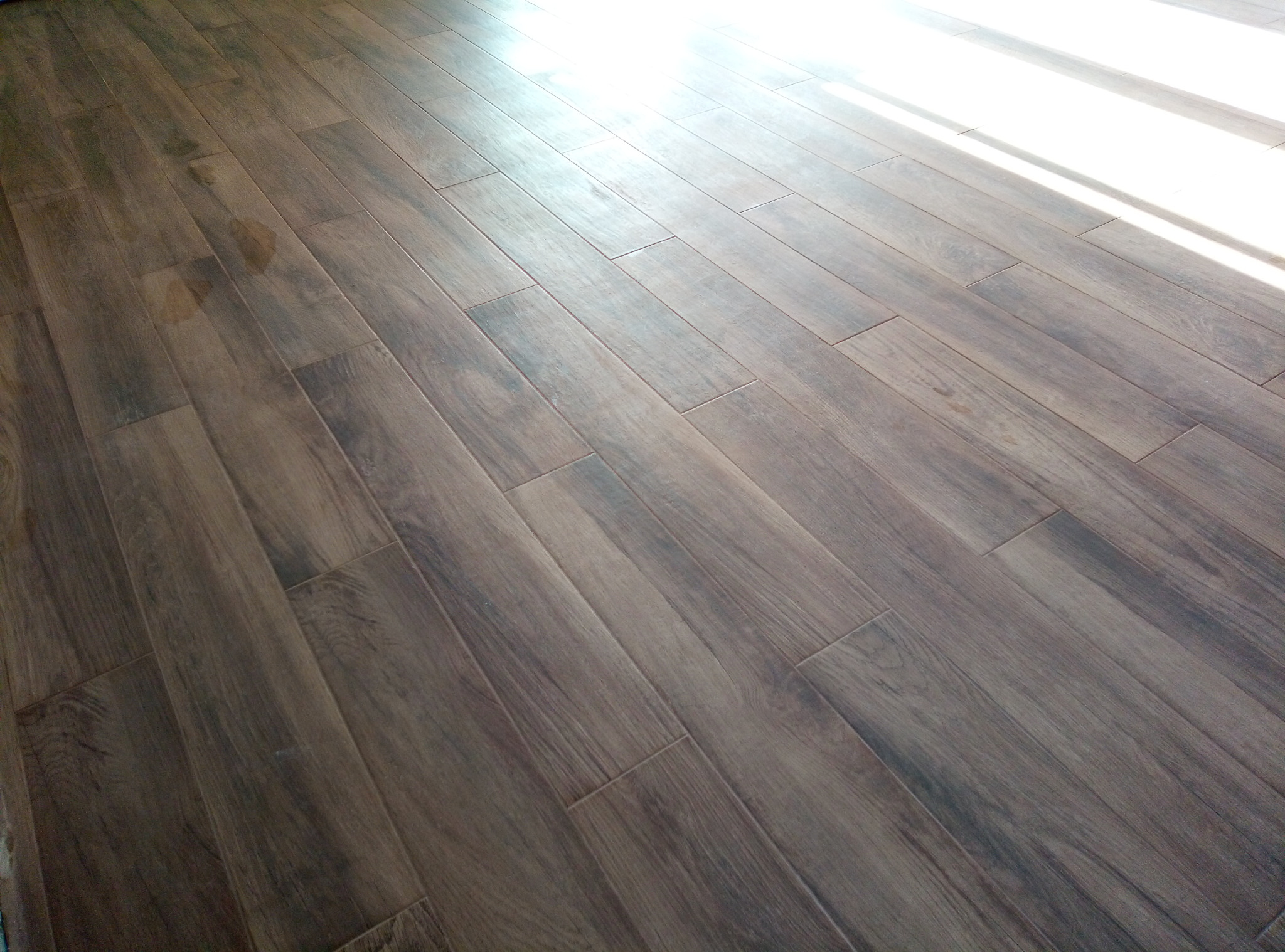 Ułożone panele podłogowe w odcieniach brązu z widocznym śladem/plamą w lewej części kadru, oświetlone naturalnym światłem wpadającym z góry po prawej stronie.