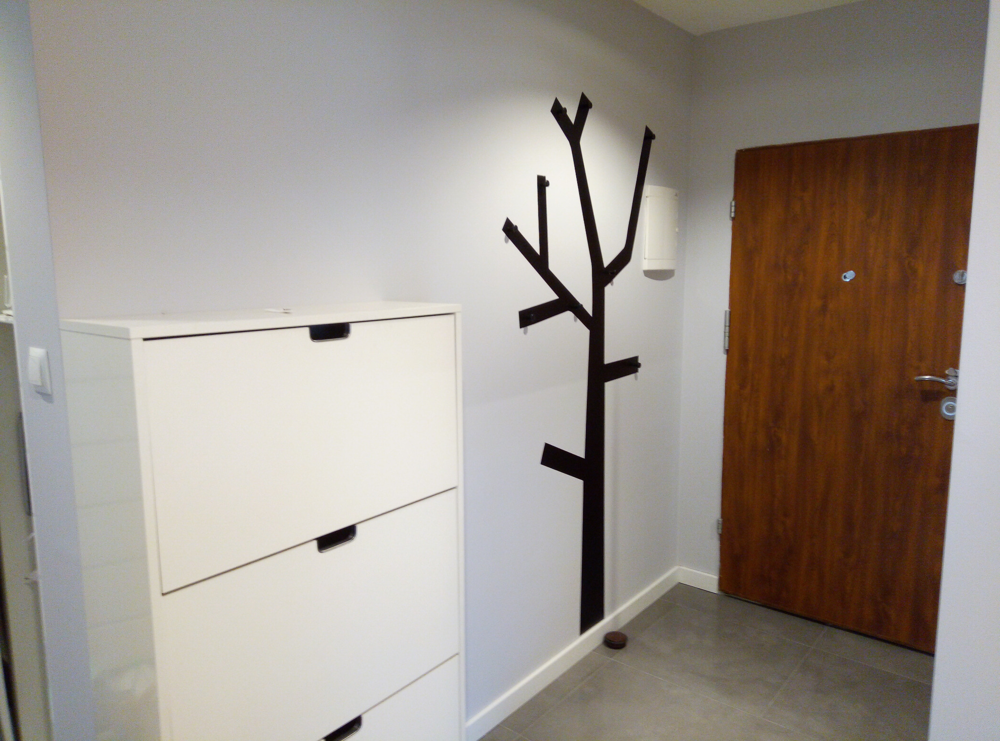 Nowoczesne wnętrze przedpokoju z białą szafką na buty, dekoracyjnym wieszakiem w kształcie drzewa i drewnianymi drzwiami wejściowymi.