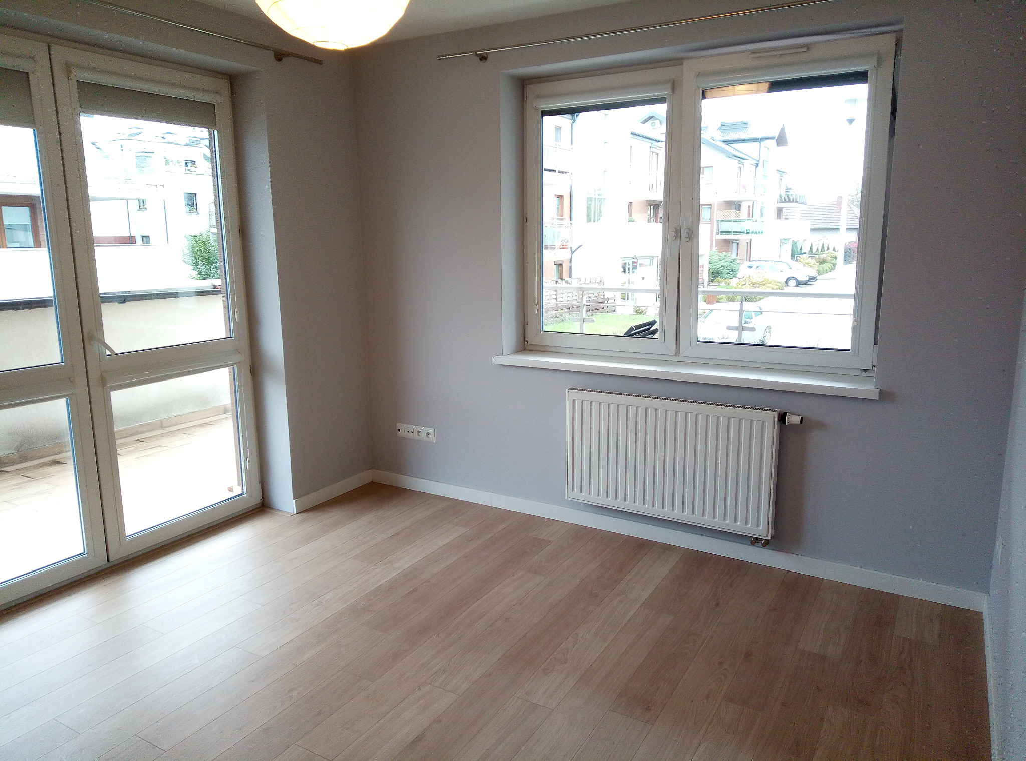 Jasny pokój z drewnianą podłogą, białymi oknami balkonowymi i grzejnikiem, ściany pomalowane na szaro, widok na budynki z zewnątrz.