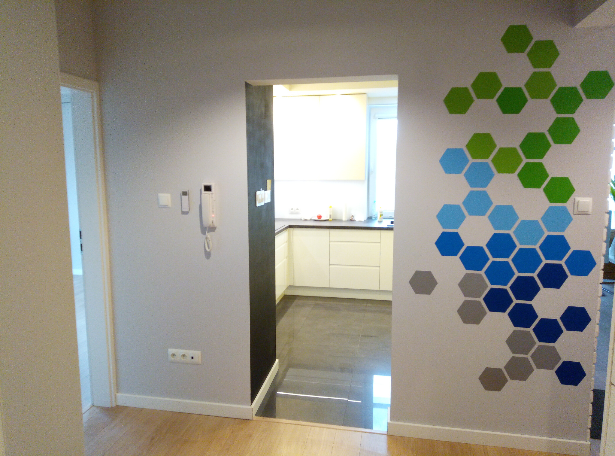 Wnętrze mieszkania z widokiem na kuchnię przez otwarte drzwi, ściana ozdobiona geometrycznym wzorem z sześciokątów w odcieniach zieleni, błękitu i szarości.