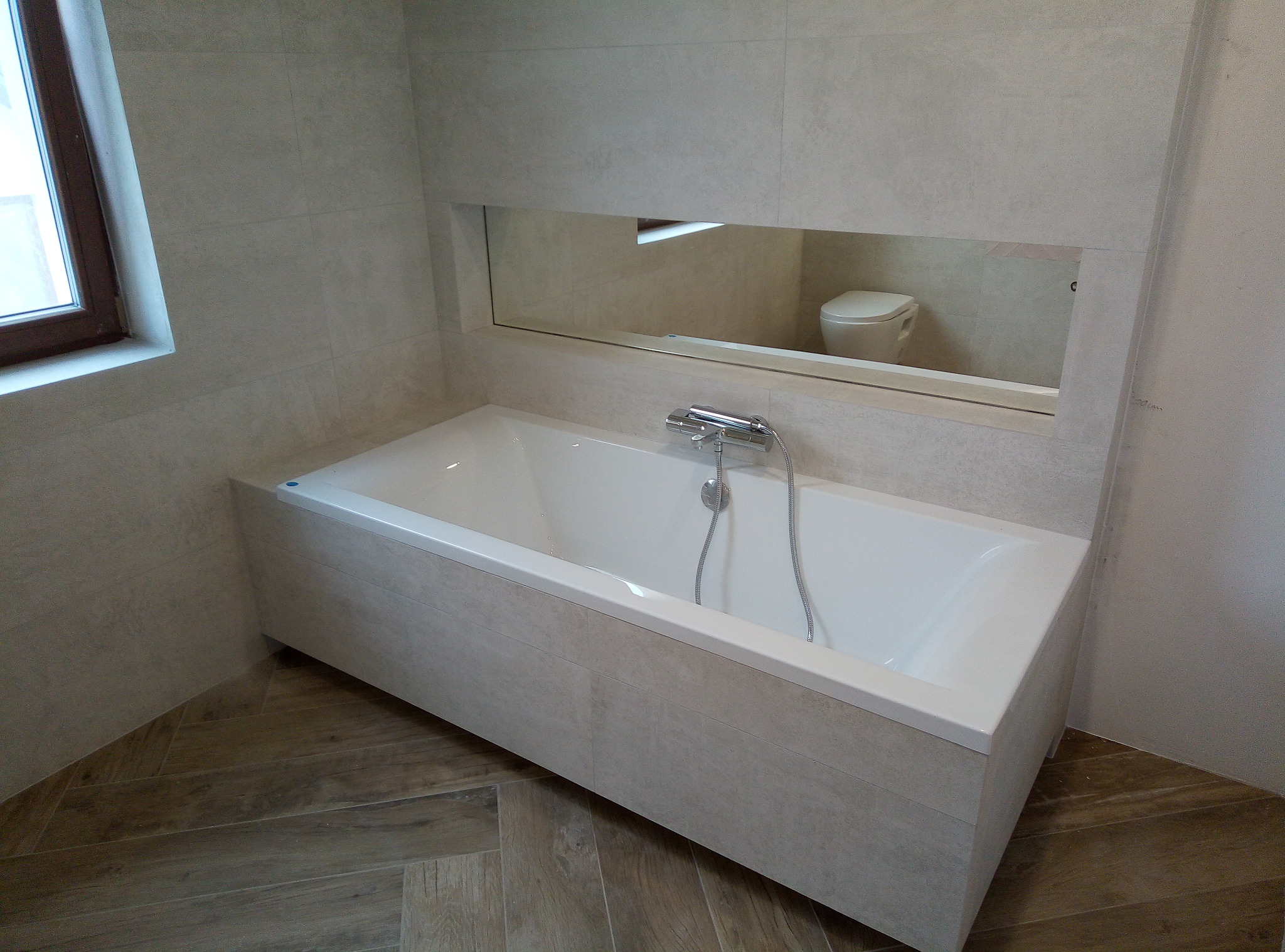 Nowoczesna łazienka z wanną obudowaną płytkami w kolorze ścian, drewnopodobna podłoga ułożona w jodełkę, w lustrze odbija się toaleta.