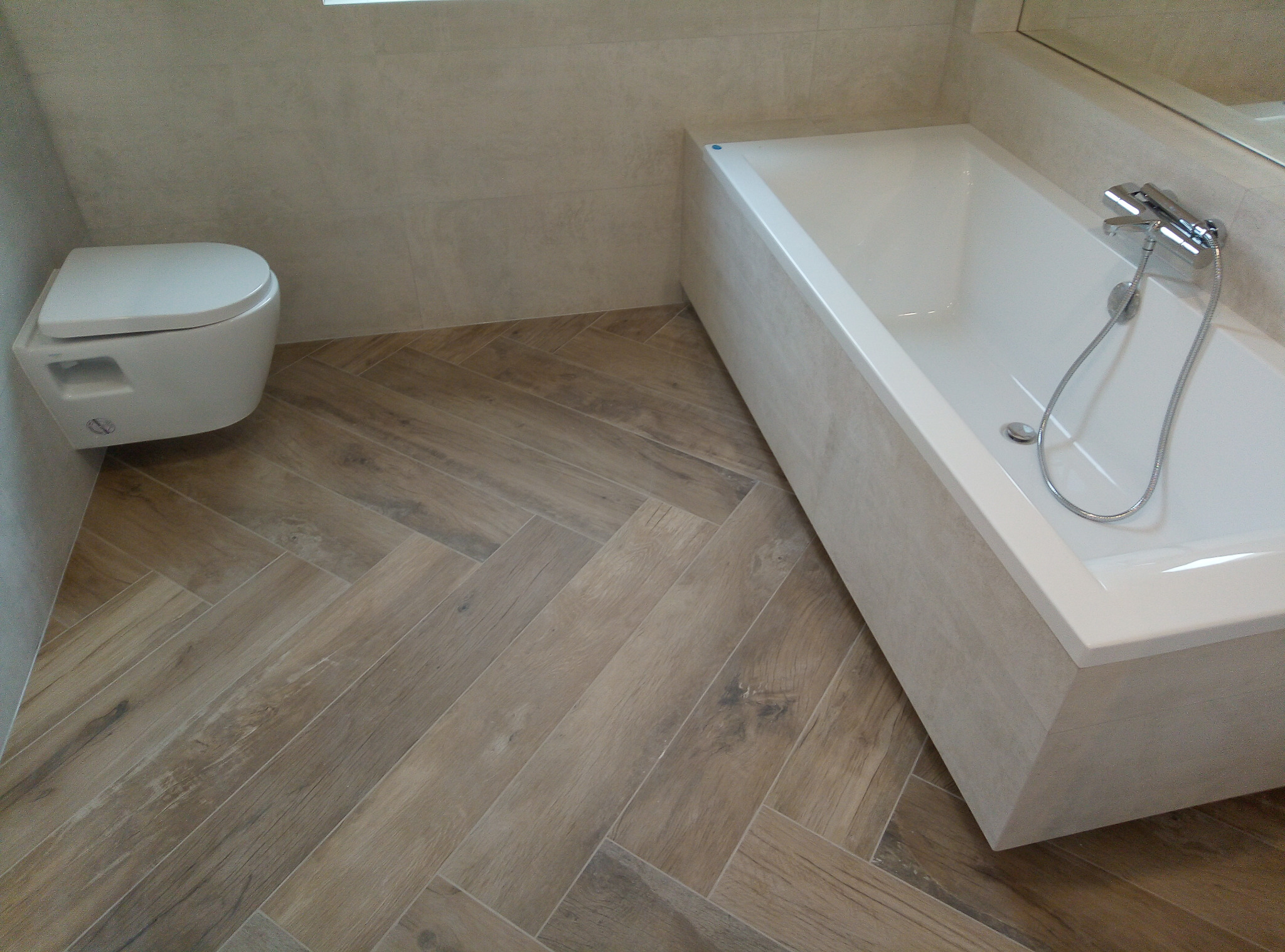 Nowoczesna łazienka z podwieszaną toaletą i wanną obudowaną płytkami w kolorze podłogi ułożonej w jodełkę.