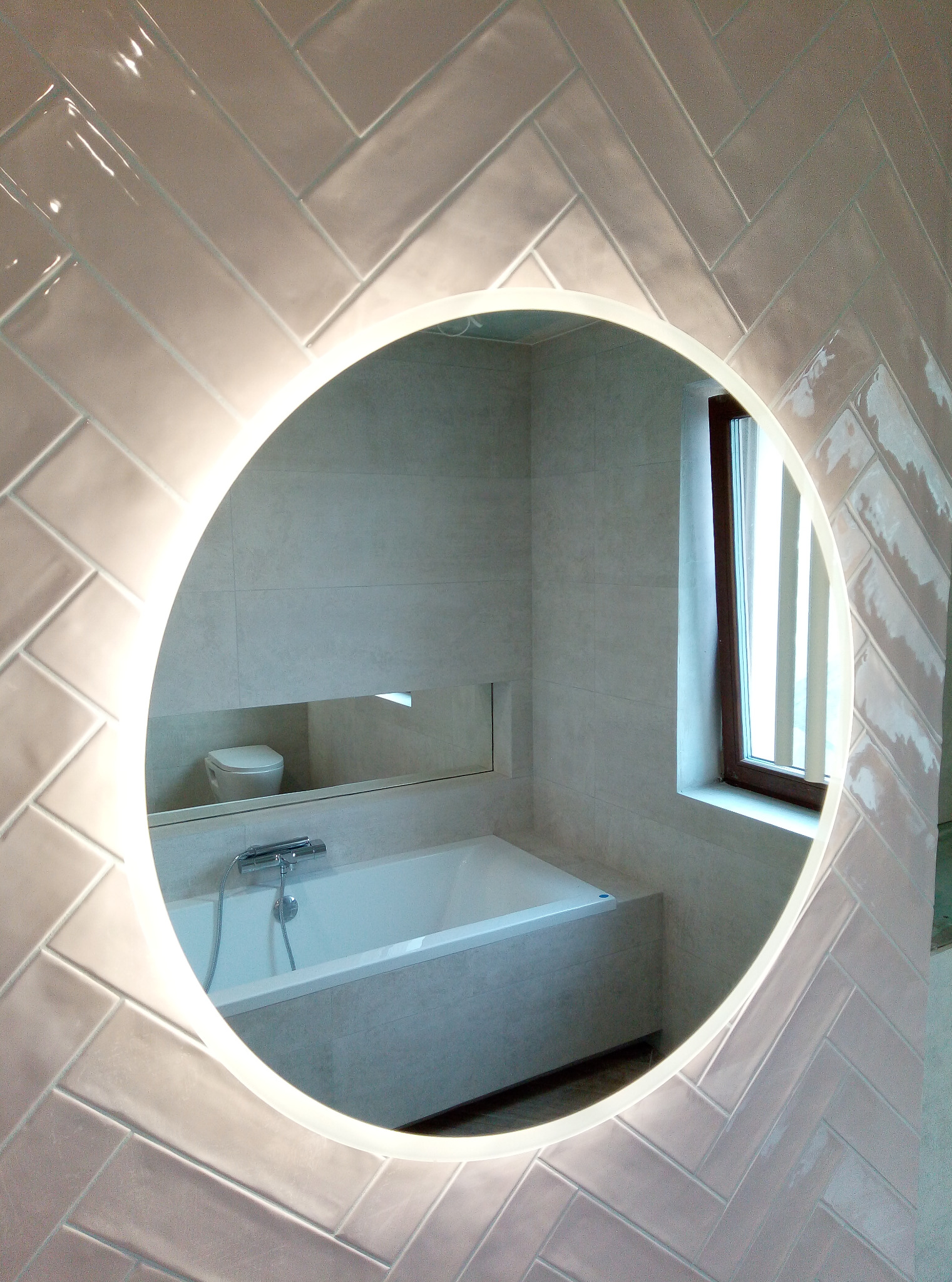 Okrągłe lustro z podświetleniem LED, odbijające wnętrze łazienki z wanną, toaletą i oknem, otoczone ścianą z ułożonych w jodełkę, błyszczących, beżowych płytek.