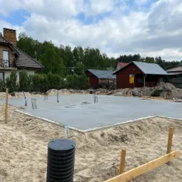 Świeżo wylana płyta fundamentowa z wystającymi prętami zbrojeniowymi, otoczona piaskiem i deskami szalunkowymi. W tle dom jednorodzinny z brązową dachówką i małym balkonem.