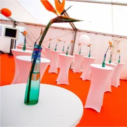 Uroczyste ustawienie stoł&oacute;w z białymi obrusami i centralnymi dekoracjami kwiatowymi w smukłych, turkusowych wazonach, w namiocie z pomarańczową podłogą, widoczny ekran w tle.