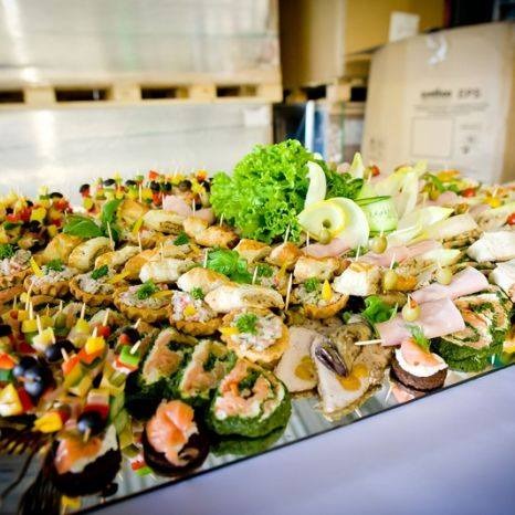 Bogato zastawiony stół z różnorodnymi kanapkami i przekąskami na srebrnej tacy, widoczne szaszłyki owocowo-warzywne, tartaletki z pastami, roladki z łososiem i szpinakiem, kanapki z szynką i serem,...