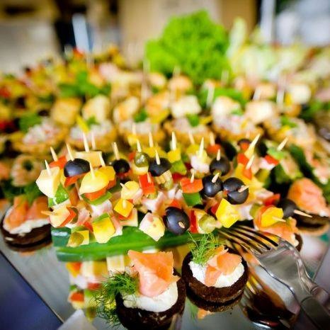 Różnorodne kolorowe przekąski na przyjęcie, w tym koreczki z serem, oliwkami i papryką oraz mini tartaletki z łososiem i koperkiem, ułożone na lustrzanej powierzchni obok srebrnych widelców.