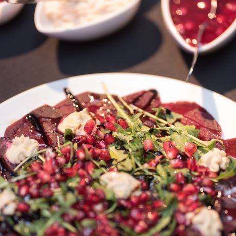 Carpaccio wołowe z rukolą, pestkami granatu i kremowym sosem, podane na białym talerzu, widoczne miseczki z dodatkowymi sosami w tle.