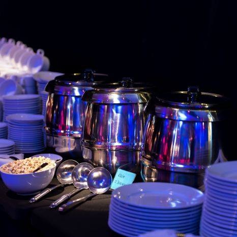 Przygotowany bufet z trzema srebrnymi bemarami, stosem białych talerzy i miseczką z przekąską, oświetlony niebieskim i różowym światłem, z widocznymi łyżkami do nakładania i karteczką z napisem.