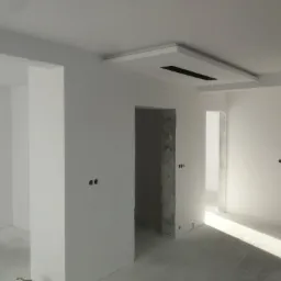 Wnętrze w trakcie białego malowania, widoczne ściany, sufit z podwieszaną konstrukcją z otworem, instalacje elektryczne w ścianach i surowy beton wok&oacute;ł ościeżnic drzwiowych.